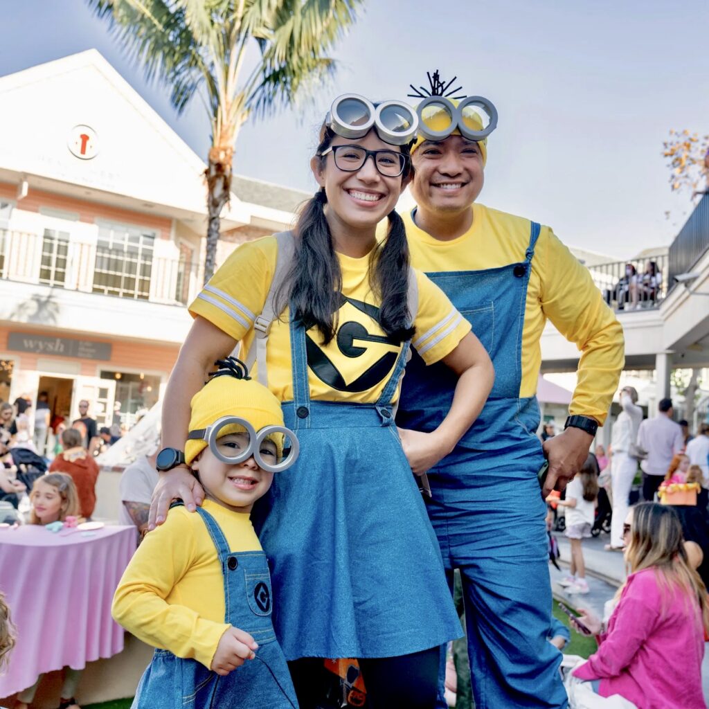A smiling family dressed in coordinated Minion costumes poses for a photo during a Halloween event at Village Faire in Carlsbad. The parents and young child wear yellow shirts, denim overalls, and Minion goggles, standing in a festive outdoor courtyard filled with other families and children in costume. The image captures the spirit of family-friendly fun and creativity at the annual Halloween Costume Contest.