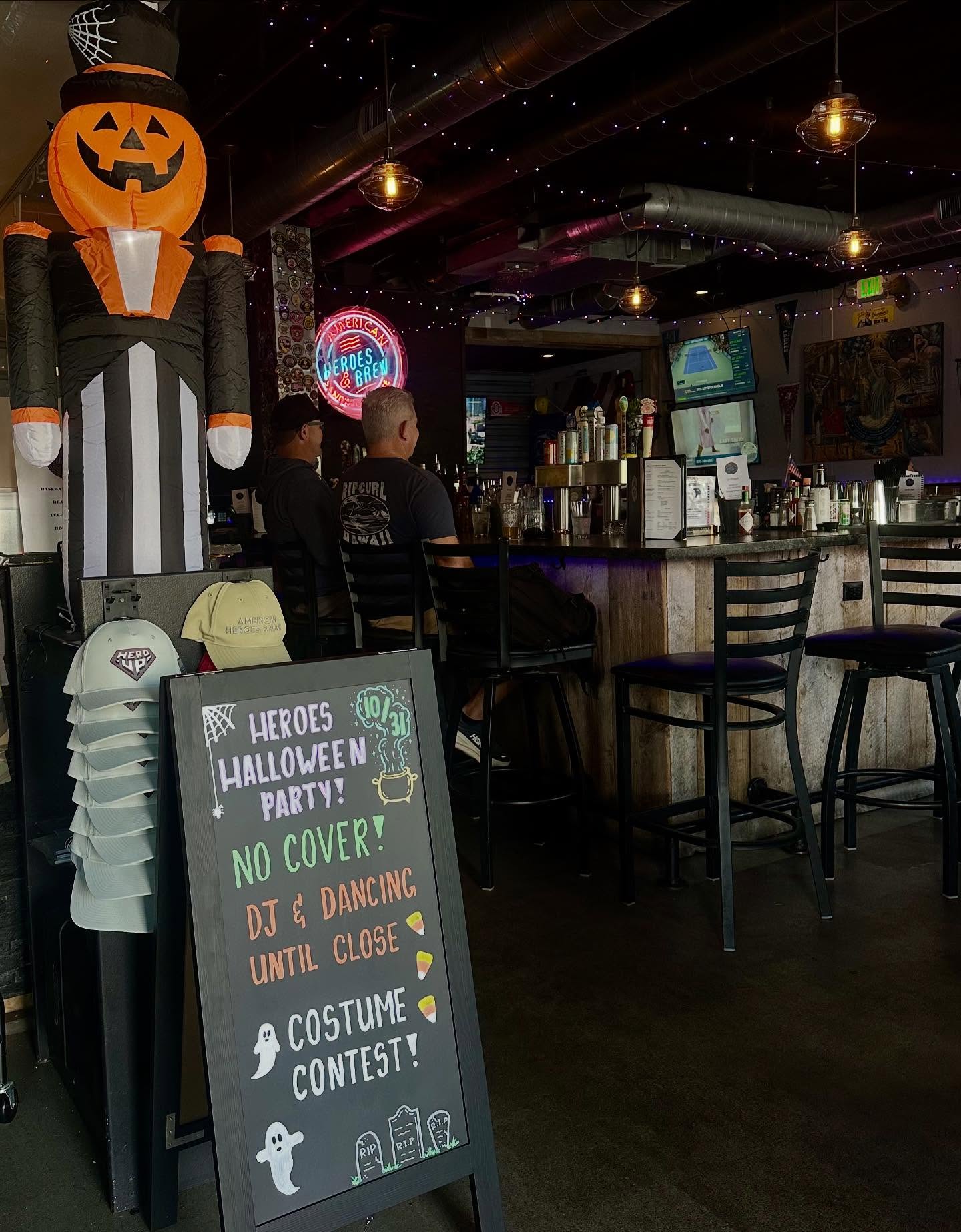 Interior of American Heroes & Brew decorated for Halloween with a large inflatable pumpkin figure, neon signage, and a chalkboard promoting the Halloween Party with DJ, dancing, and costume contest.